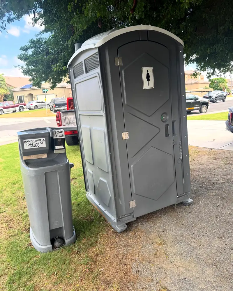 Grey standard porta potty with a freestanding handwash sink on grass